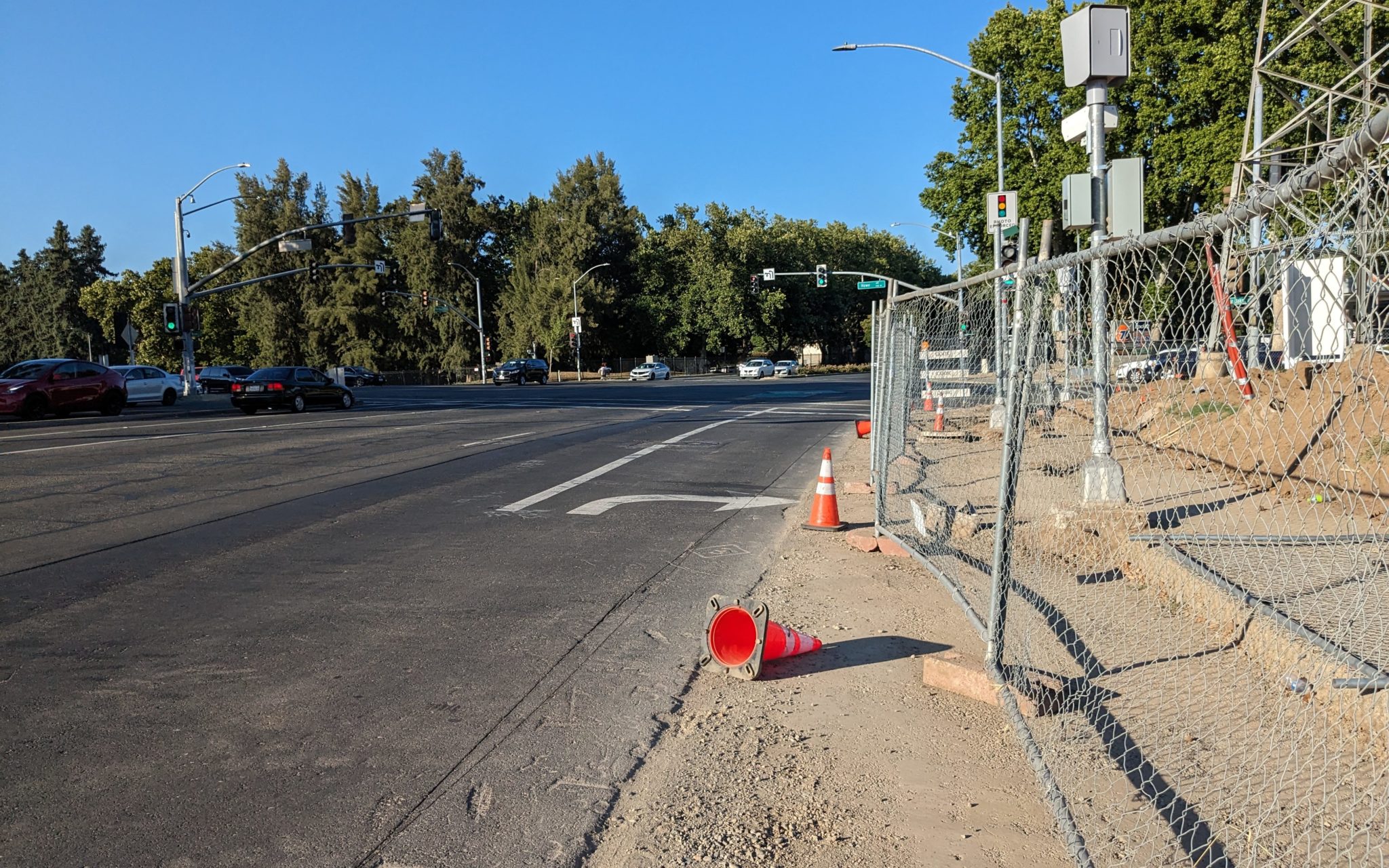 Stroad with orange traffic cones nearby and a cyclone fence around the sidewalk. One traffic cone is knocked over. 