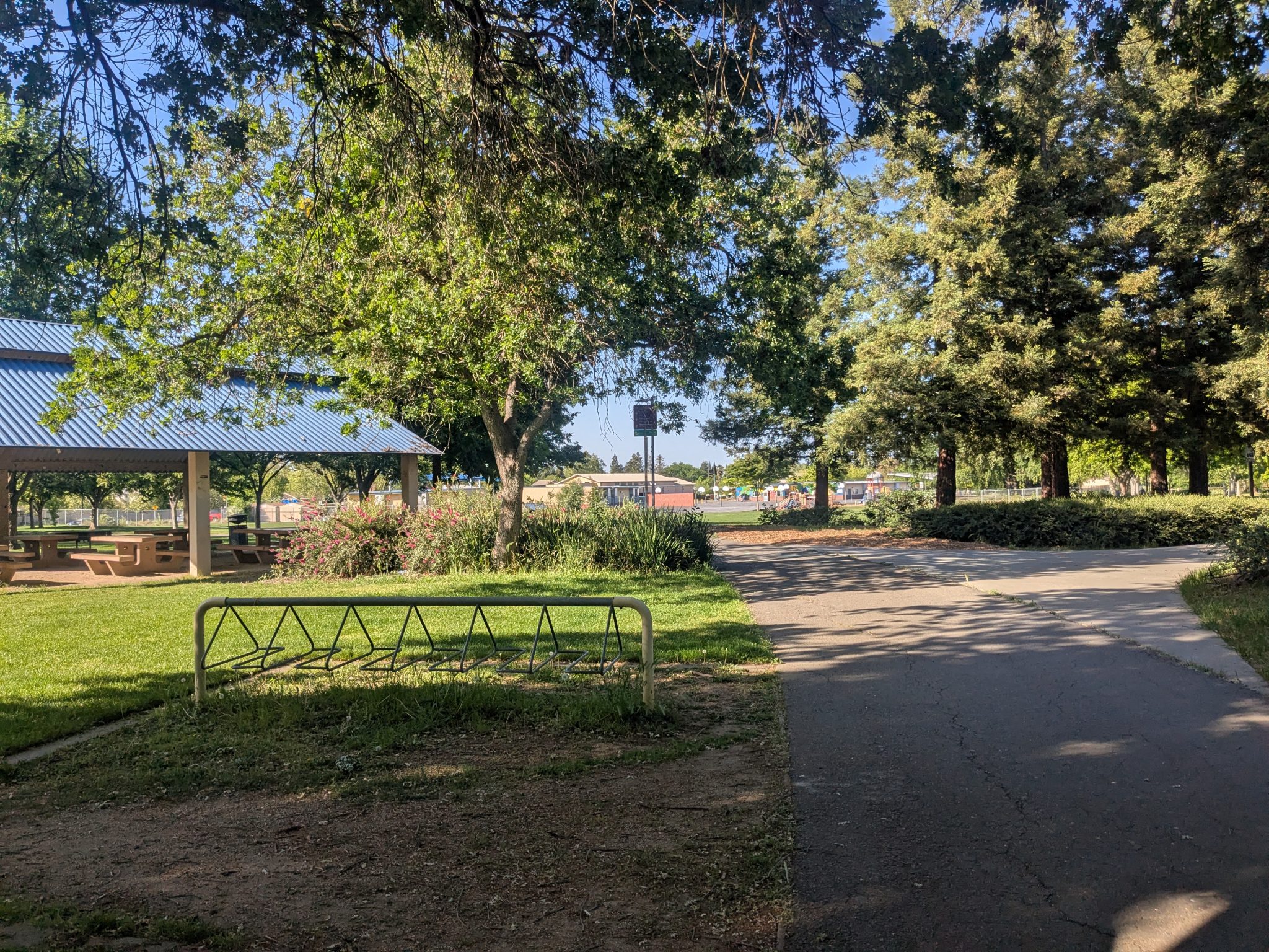 Bicycle racks near path with green grass and trees around. Covered seating area is in the back.
