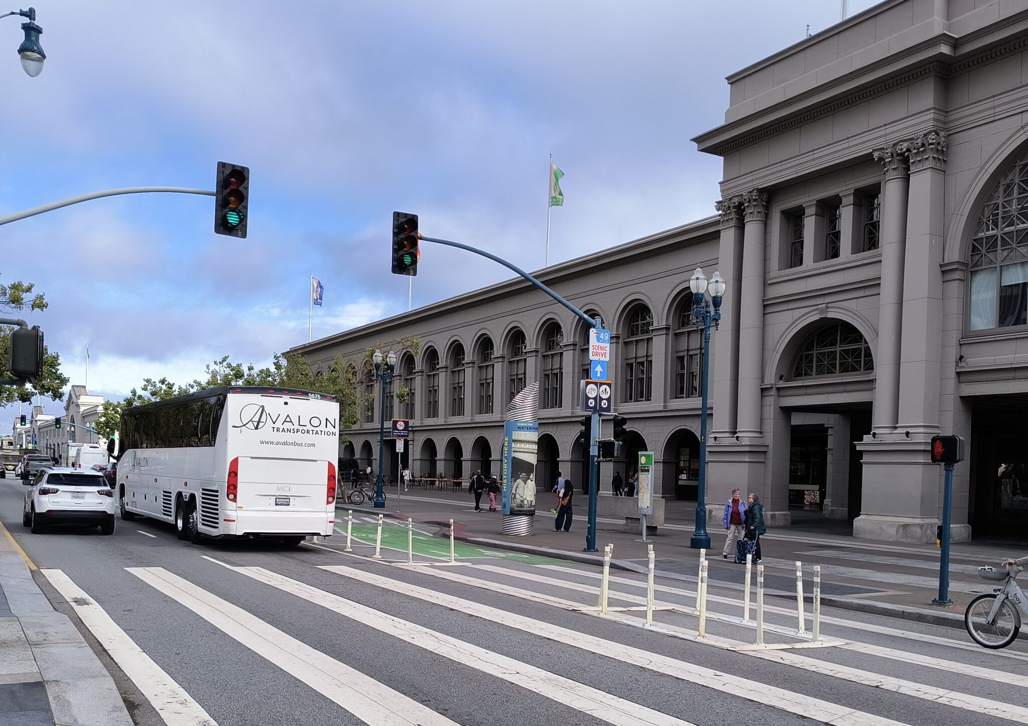 Picture of a street with cars on it. A bike lane is on the right of the street, with flex posts between the cars and bike path. People are walking on the sidewalk in front of the SF Ferry building. 